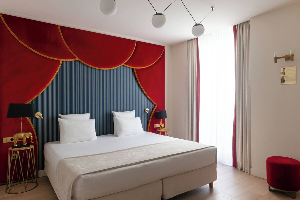 Habitación de hotel con una cama grande, dos mesitas de noche con lámparas, una pared de acento roja y una ventana con cortinas.