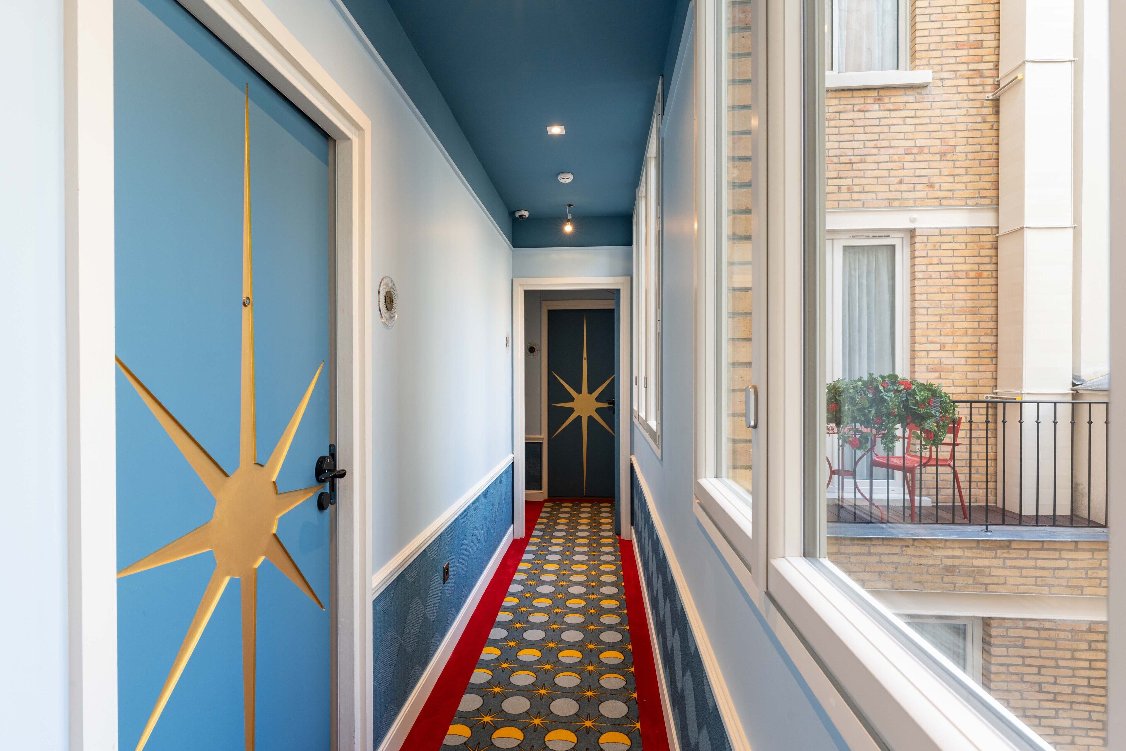 A narrow hallway with a star-shaped door, a patterned red and yellow carpet, and a view of a balcony with a brick building and a table with chairs.