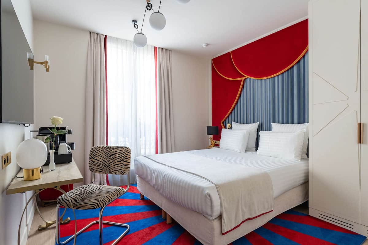 Habitación de hotel moderna con una cama grande, un escritorio con lámpara, una silla y una alfombra a rayas azules y rojas.