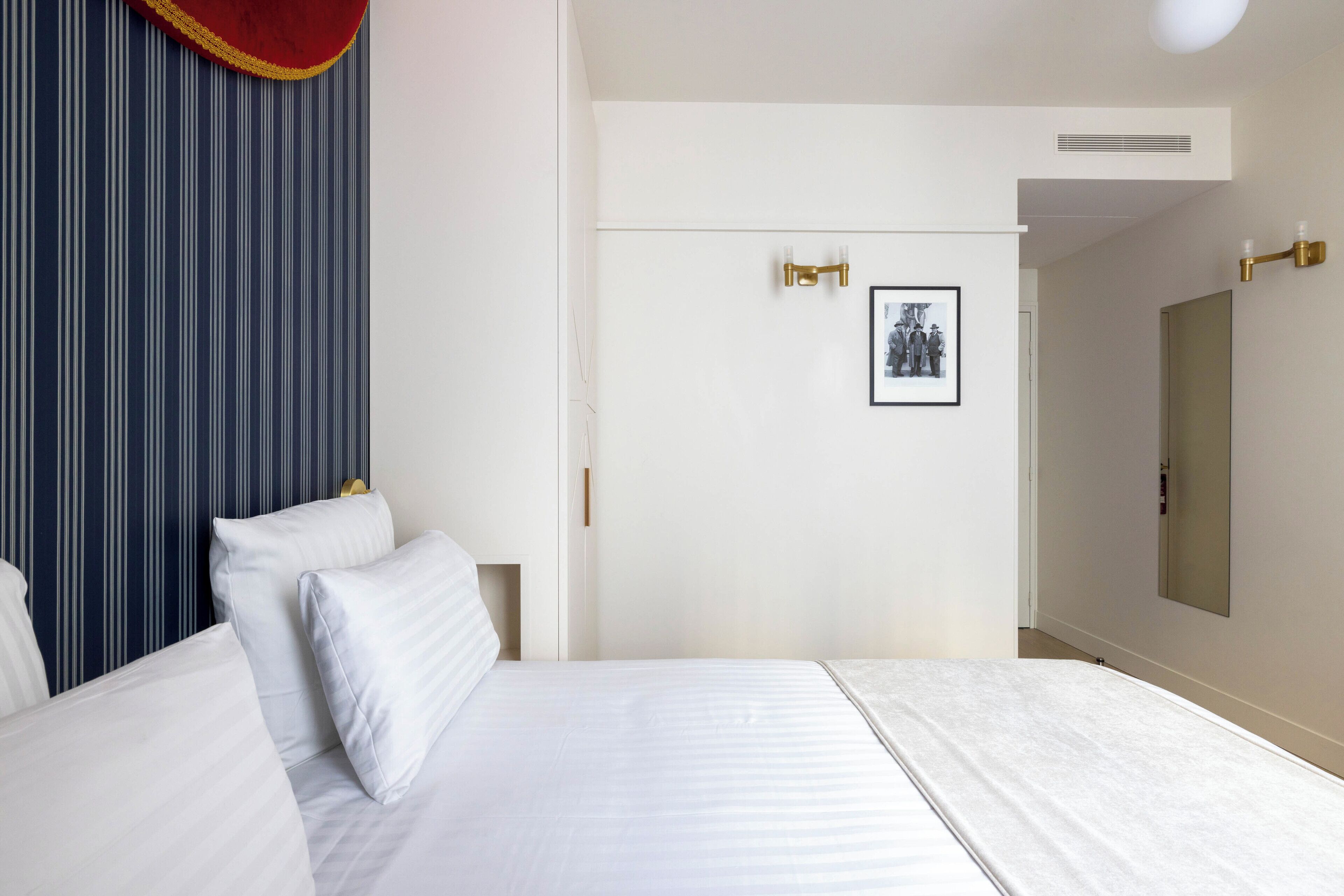 A hotel room with a large bed, a headboard with a red and gold trim, a framed picture on the wall, and a mirror on the opposite wall.