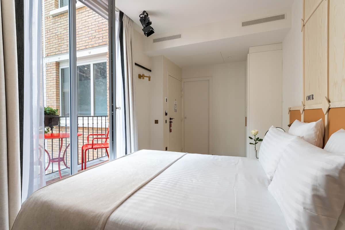 Habitación de hotel con una cama grande, un ventanal corredero que da a un área exterior con sillas rojas y vistas de un edificio de ladrillo.