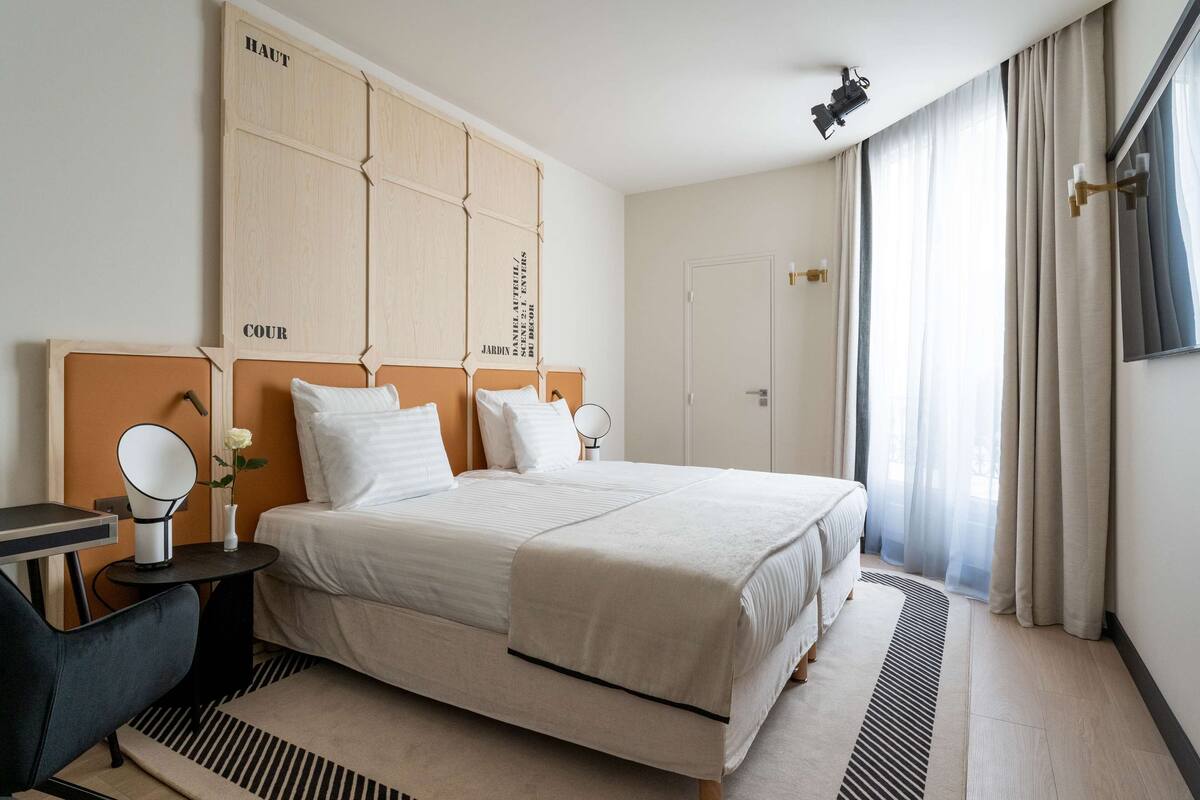 Habitación de hotel con una cama grande, una mesita de noche con lámpara, una pequeña mesa con espejo y una ventana con cortinas.