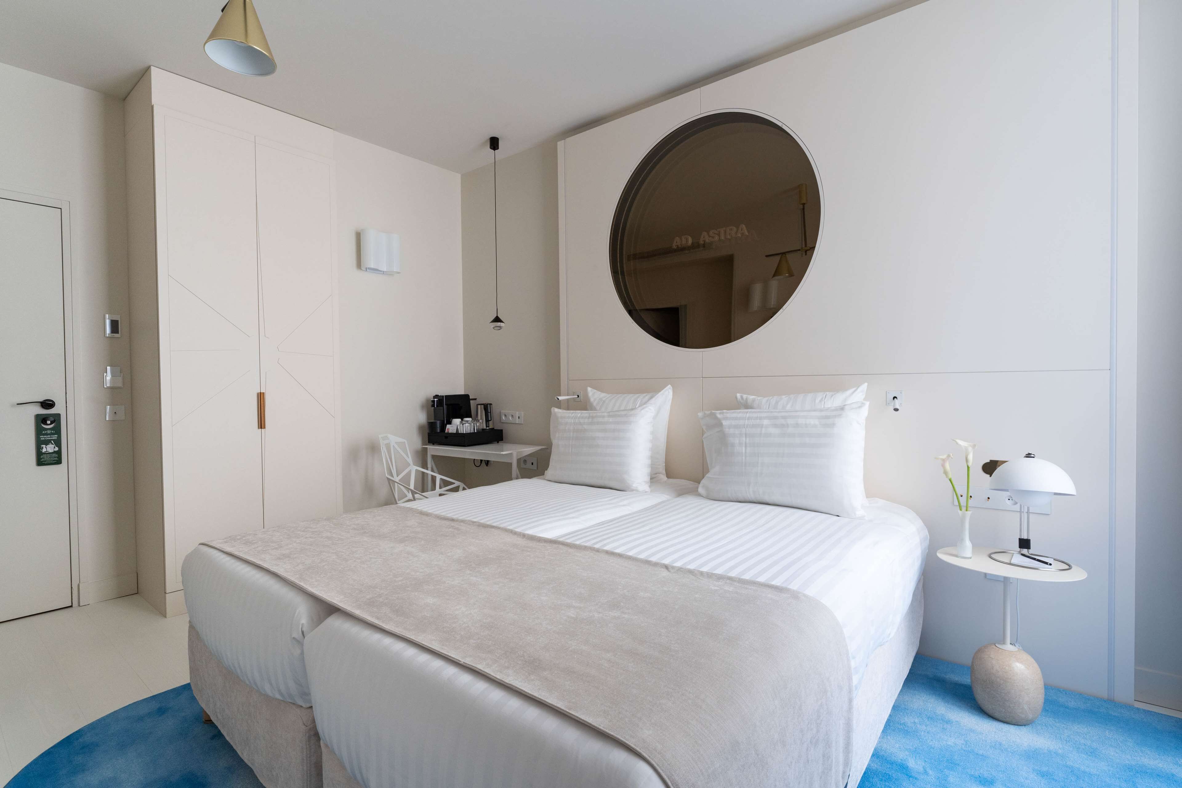Ein modernes Hotelzimmer mit einem großen Bett, einem runden Spiegel, einem kleinen Schreibtisch und einem Nachttisch mit Lampe.