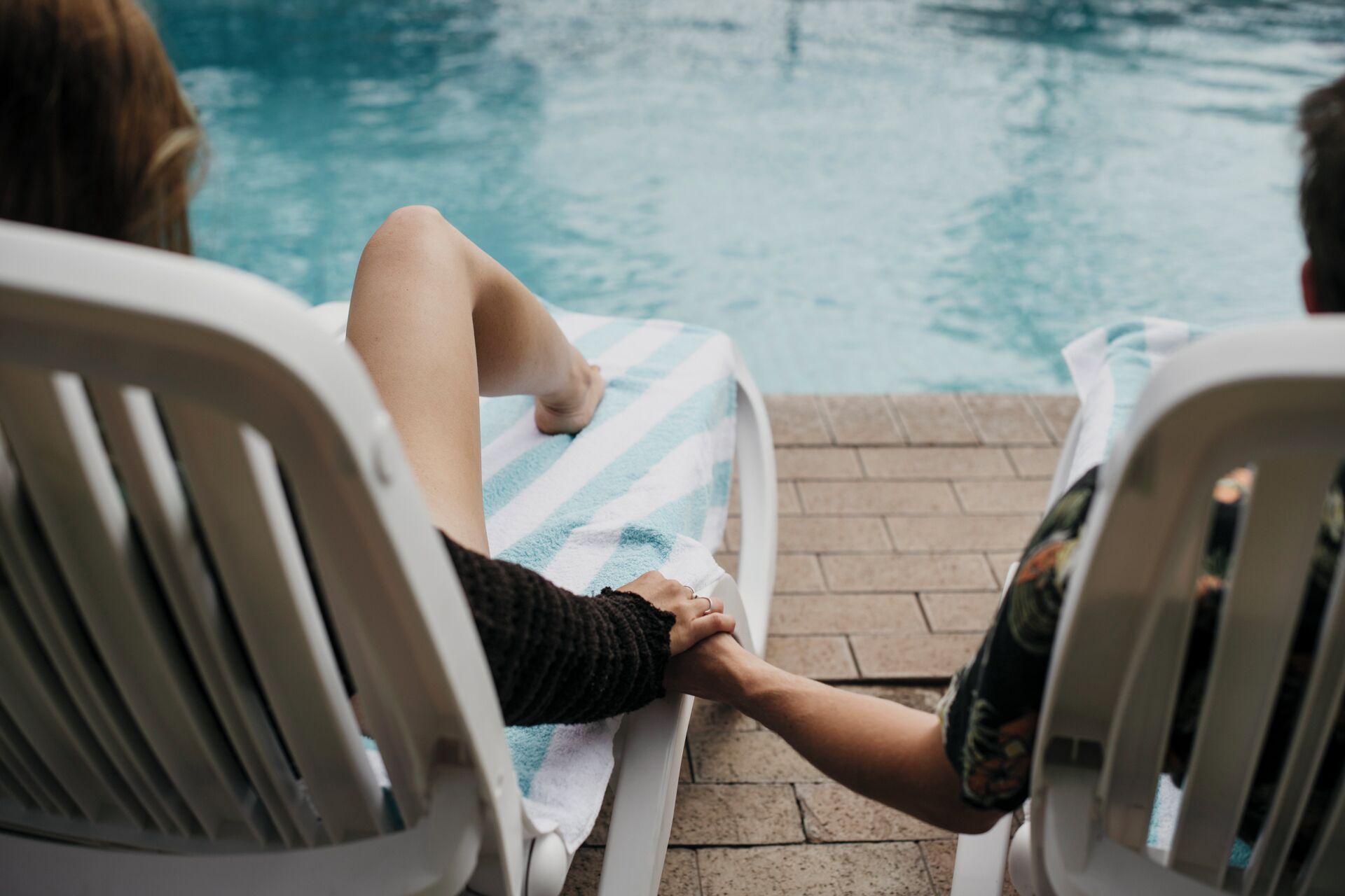 Two people sitting on lounge chairs by a pool, holding hands.