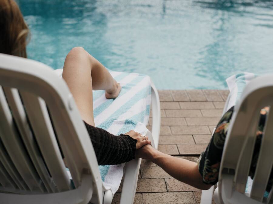 Dos personas sentadas en sillones junto a una piscina, tomadas de la mano.