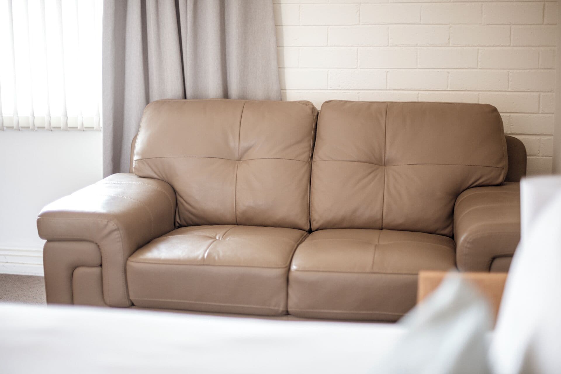 A tan leather sofa against a white brick wall with a window and curtains.