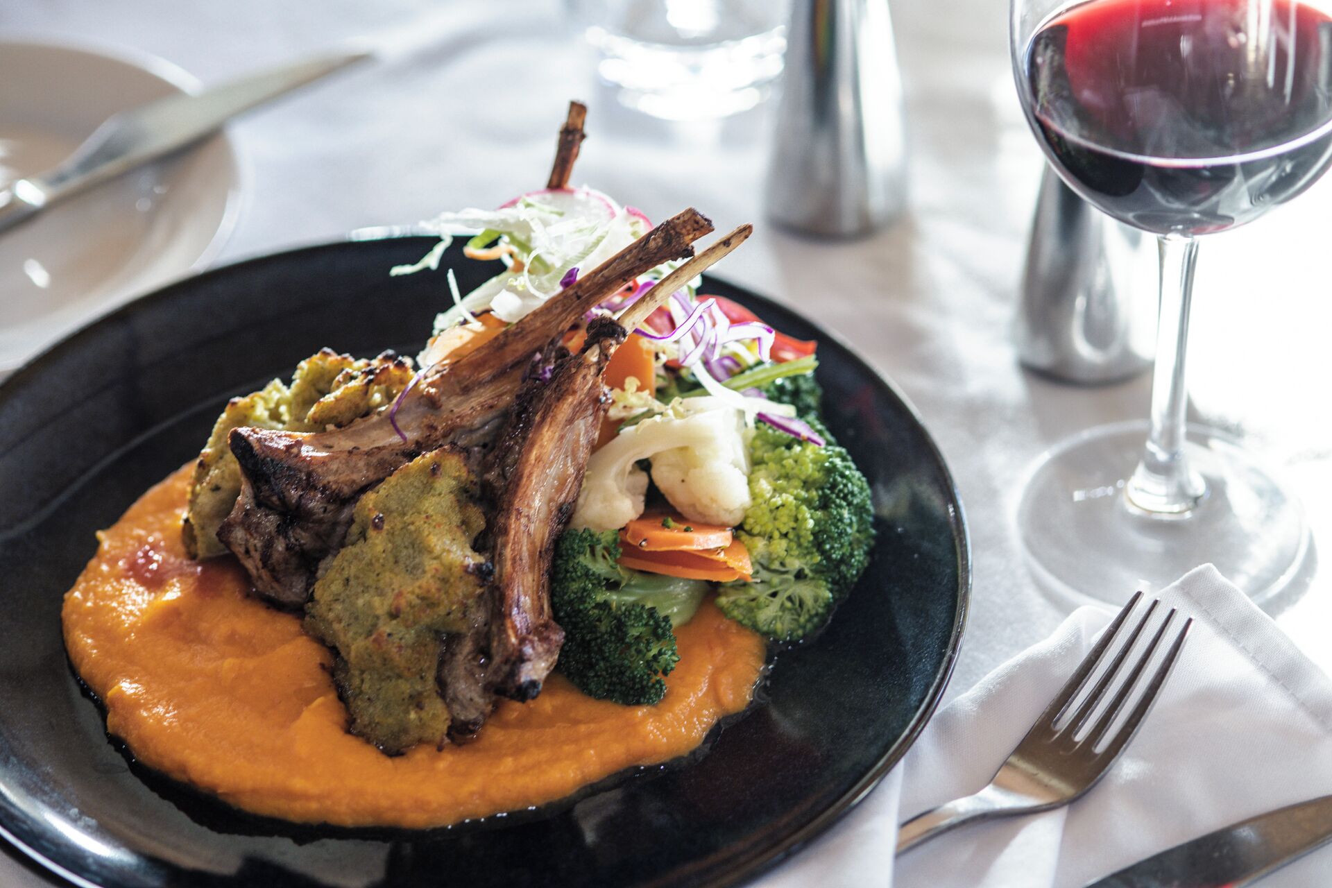 A plate of food with a lamb shank, vegetables, and a sauce, accompanied by a glass of red wine.