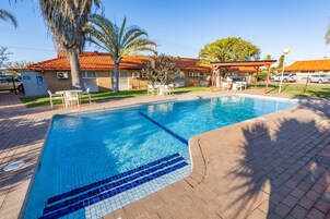 Piscina com deck azulejado, cercada por palmeiras e um edifício com telhado vermelho.