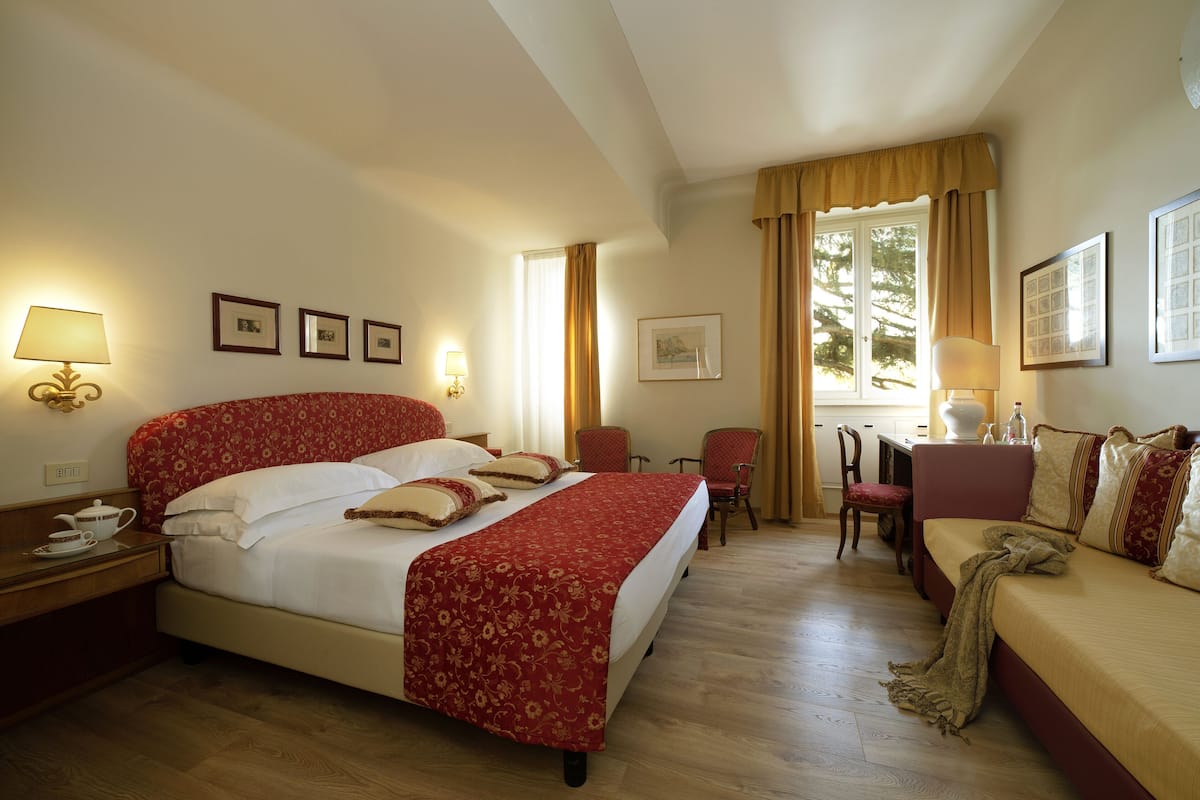 A hotel room with a large bed, a sofa, a desk, and a chair. There are framed pictures on the wall, a lamp on the desk, and a window with curtains.