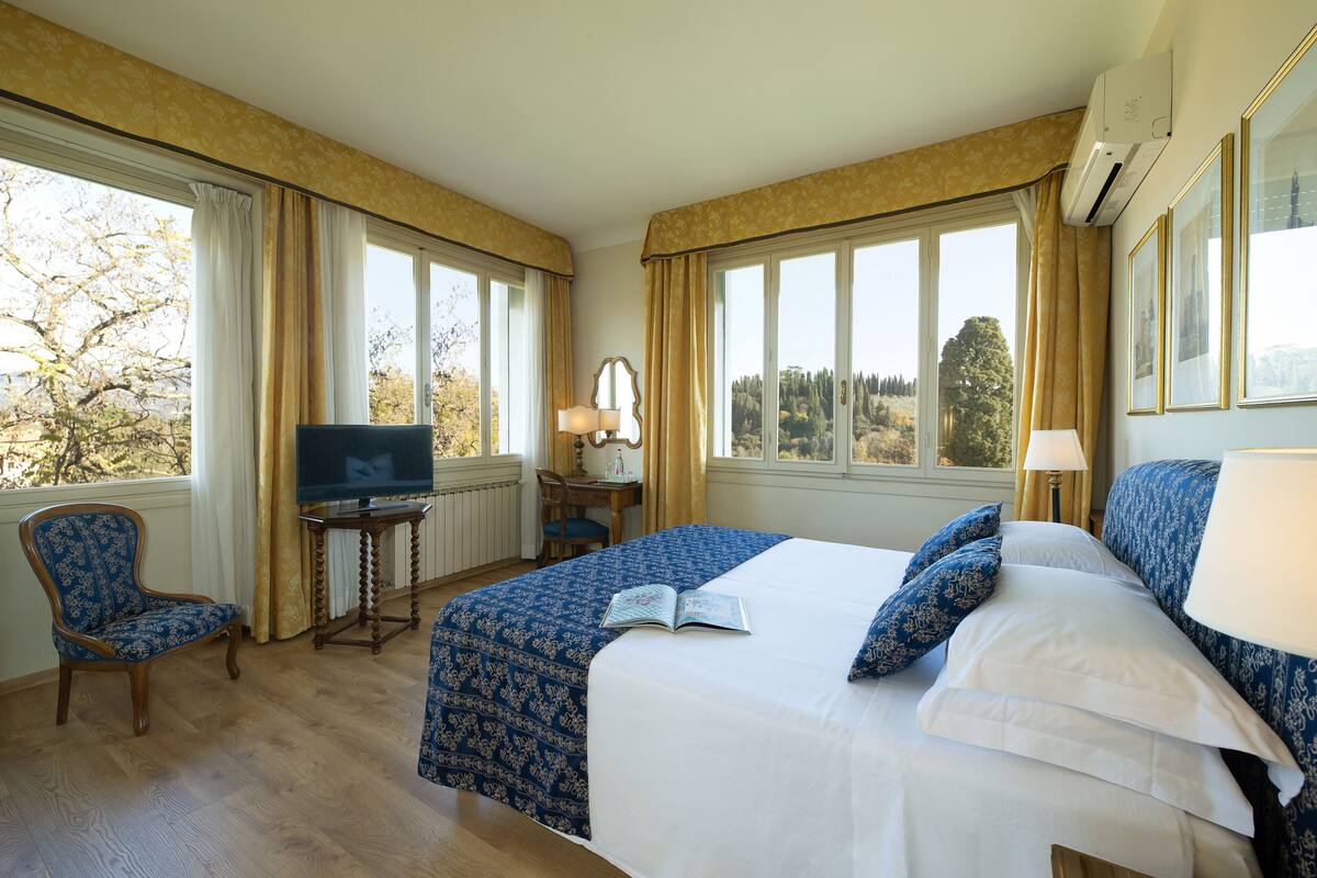 A hotel room with a large bed, a desk with a chair, a television, and large windows with curtains.