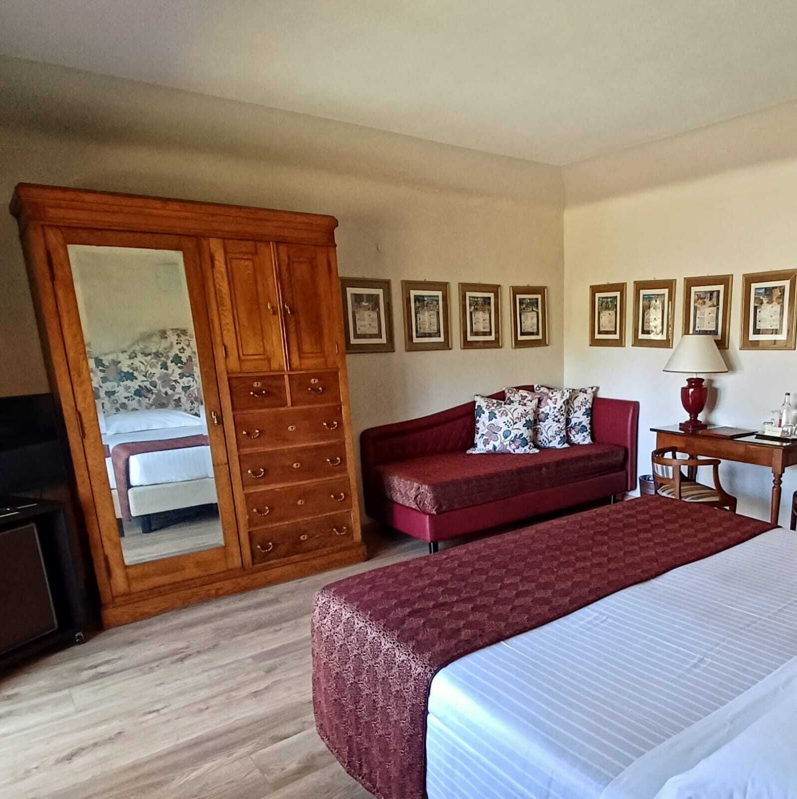 A hotel room with a wooden wardrobe, a red sofa, a bed, and a desk with a lamp.