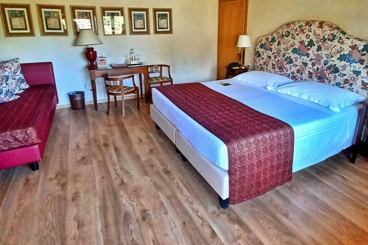 A bedroom with a bed, a desk, two chairs, a red sofa, and a wooden floor.