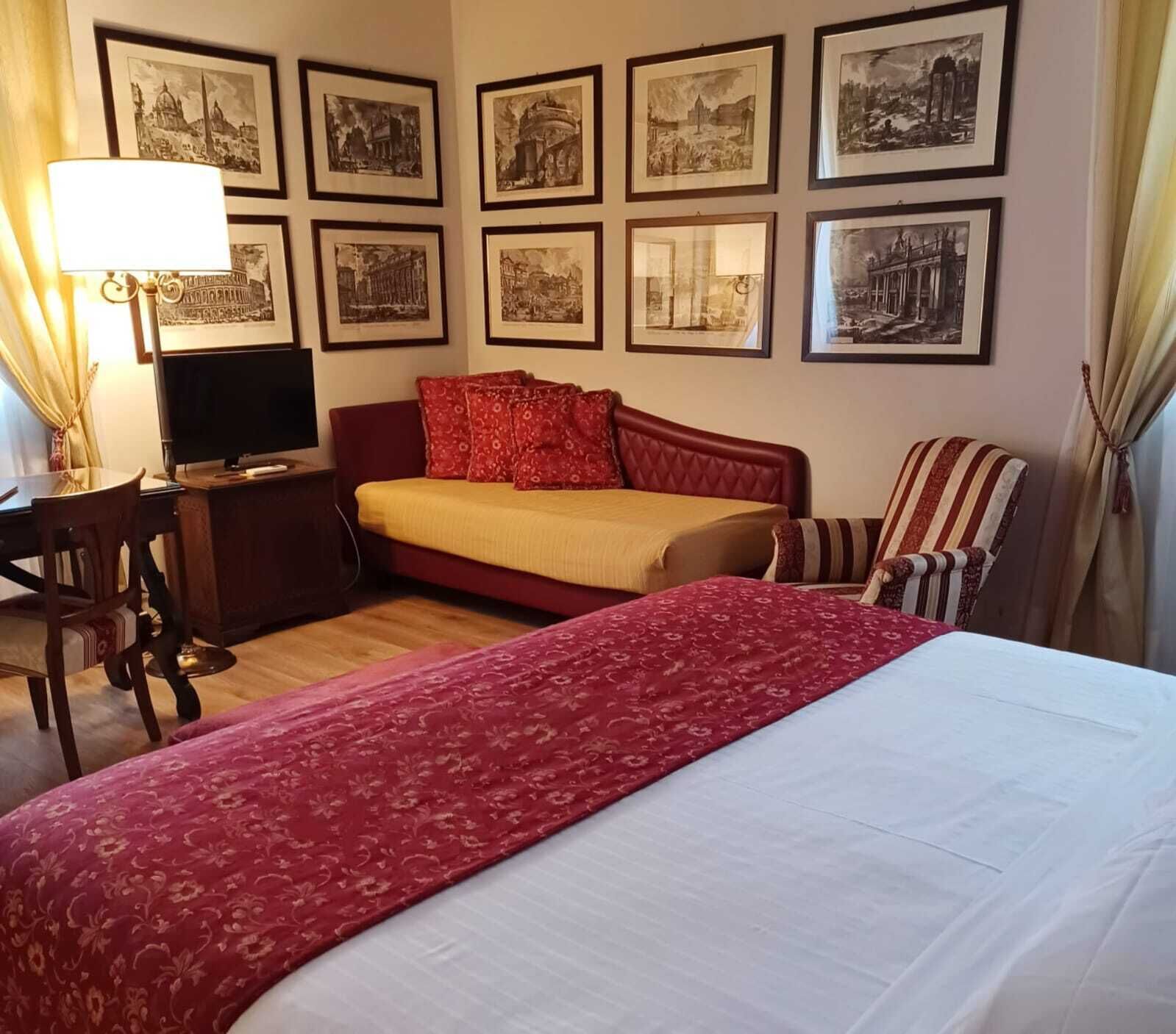A hotel room with a large bed, a sofa bed, a desk with a lamp, and a chair. There are framed pictures on the wall.