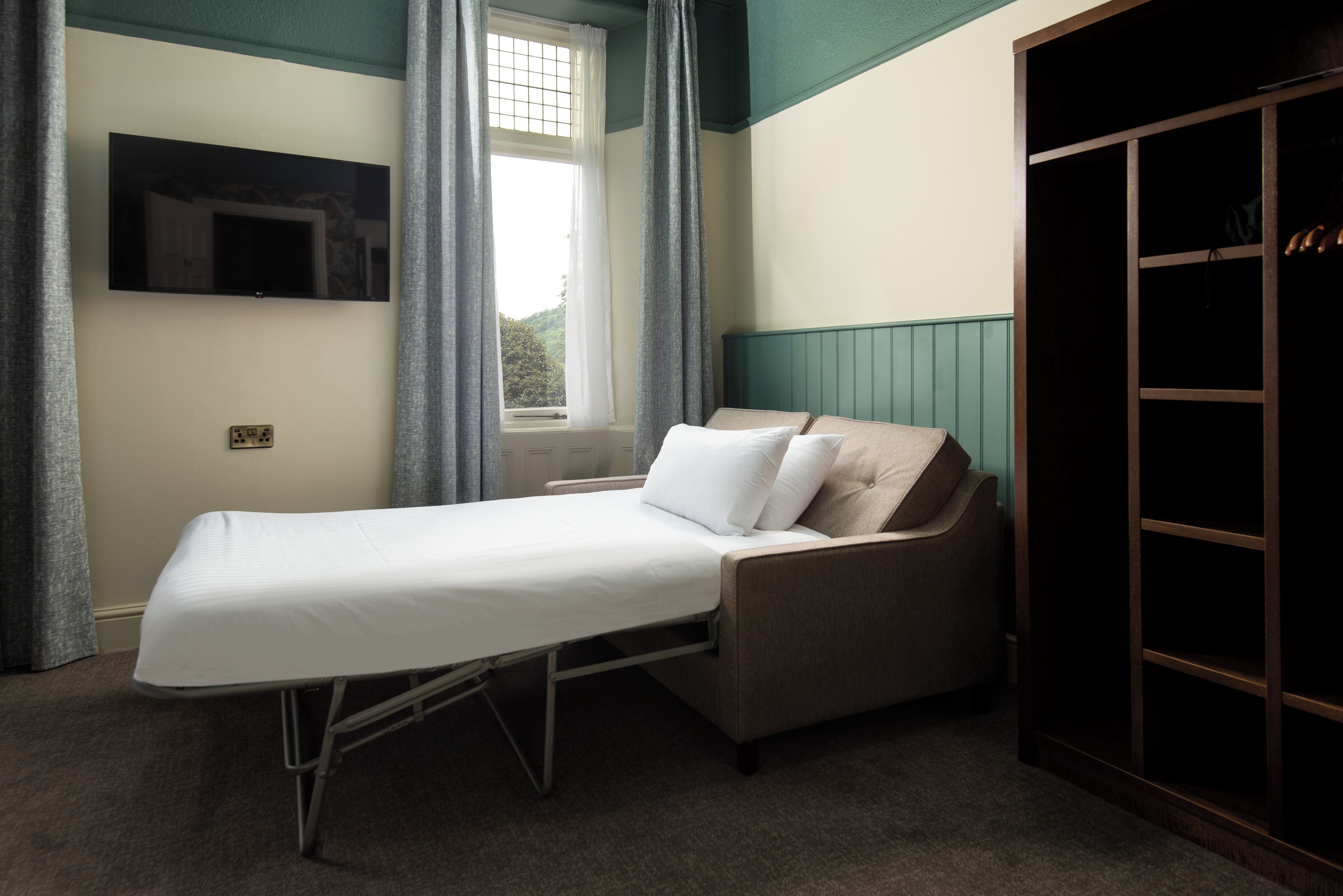 A single-bed room with a TV mounted on the wall, a window with curtains, and a wardrobe.