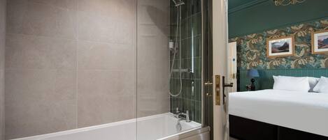 A modern bathroom with a glass-enclosed shower, a white bathtub, and a bed with a dark headboard in the adjacent room.