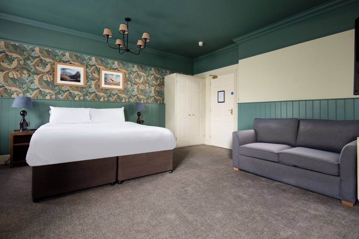 A bedroom with a bed, a gray sofa, a nightstand, and a wall with floral wallpaper.