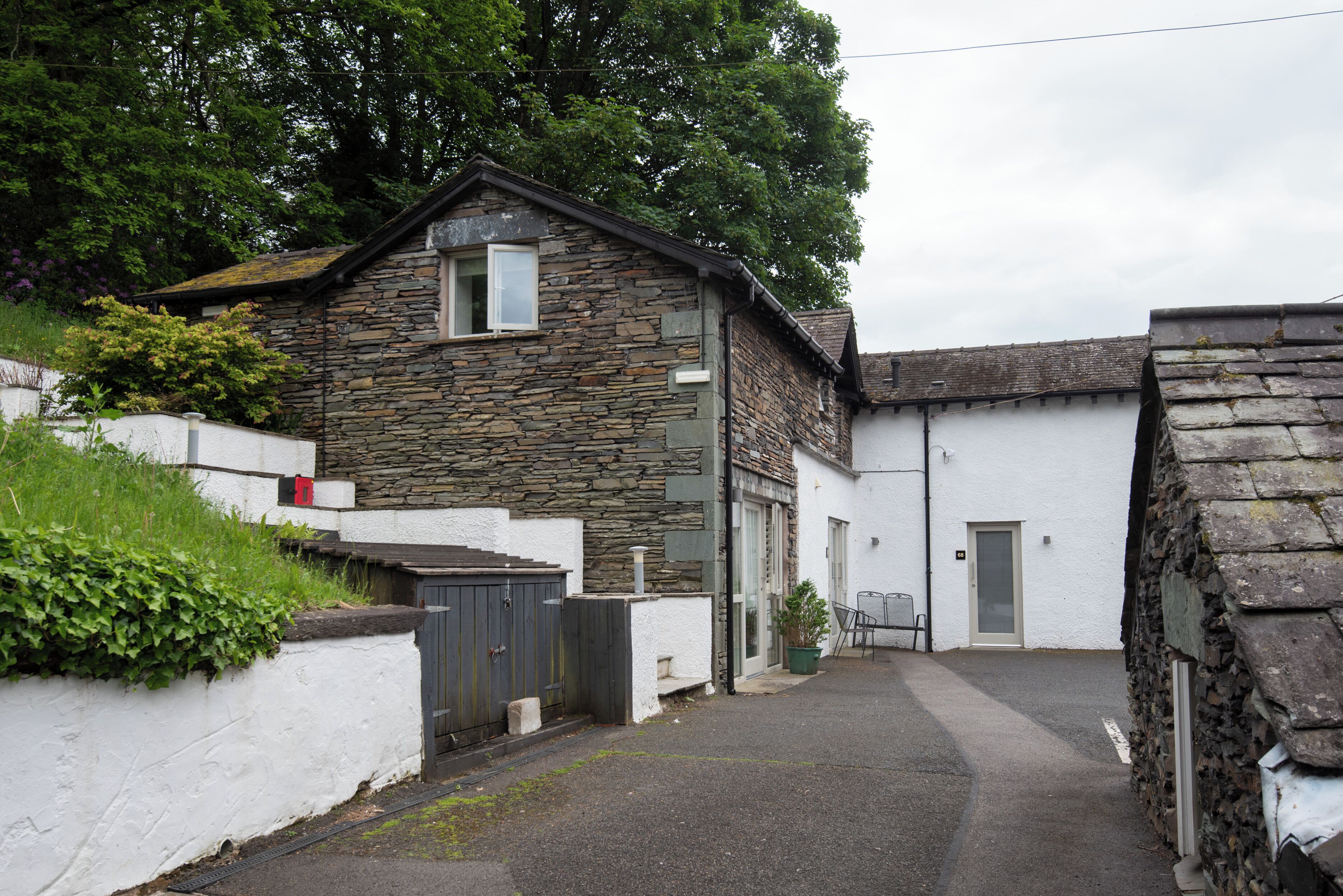 A stone building with a small window and a door, surrounded by a white wall and greenery.