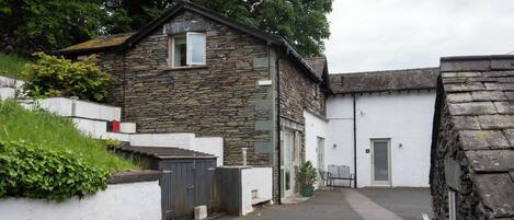 A stone building with a small window and a door, surrounded by a white wall and greenery.