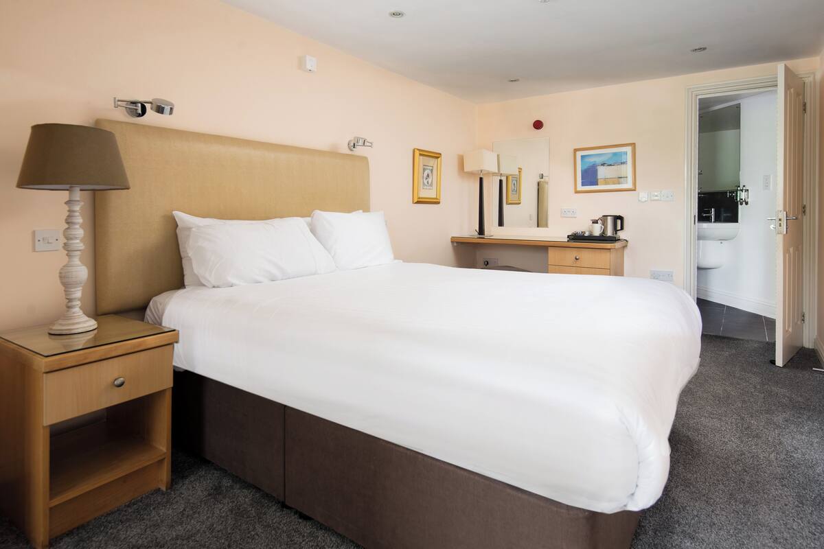 A hotel room with a large bed, a bedside table with a lamp, a dresser, a mirror, and a door leading to a bathroom.