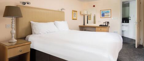 A hotel room with a large bed, a bedside table with a lamp, a dresser, a mirror, and a door leading to a bathroom.