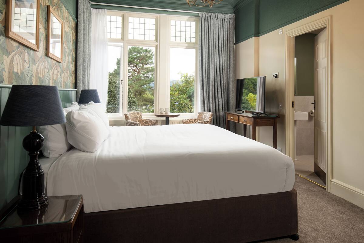 A neatly made bed with white linens, a television on a wooden stand, and a window with curtains.