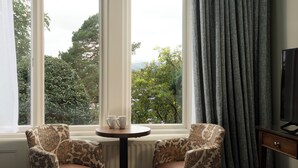 Une chambre d’hôtel avec un lit, deux fauteuils à motifs, une petite table avec deux tasses, et une vue sur la verdure grâce à de grandes fenêtres.
