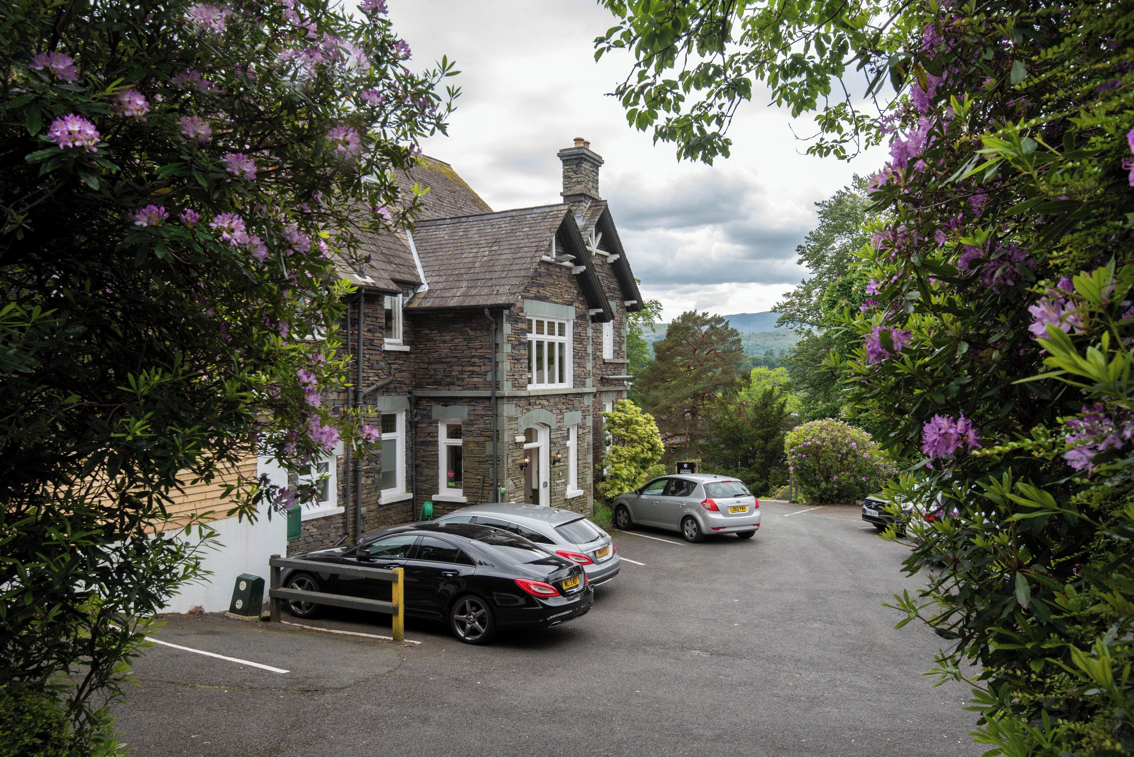 A stone house with a sloped roof and a chimney, surrounded by lush greenery and purple flowers.