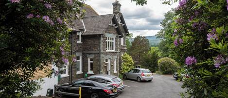 A stone house with a sloped roof and a chimney, surrounded by lush greenery and purple flowers.