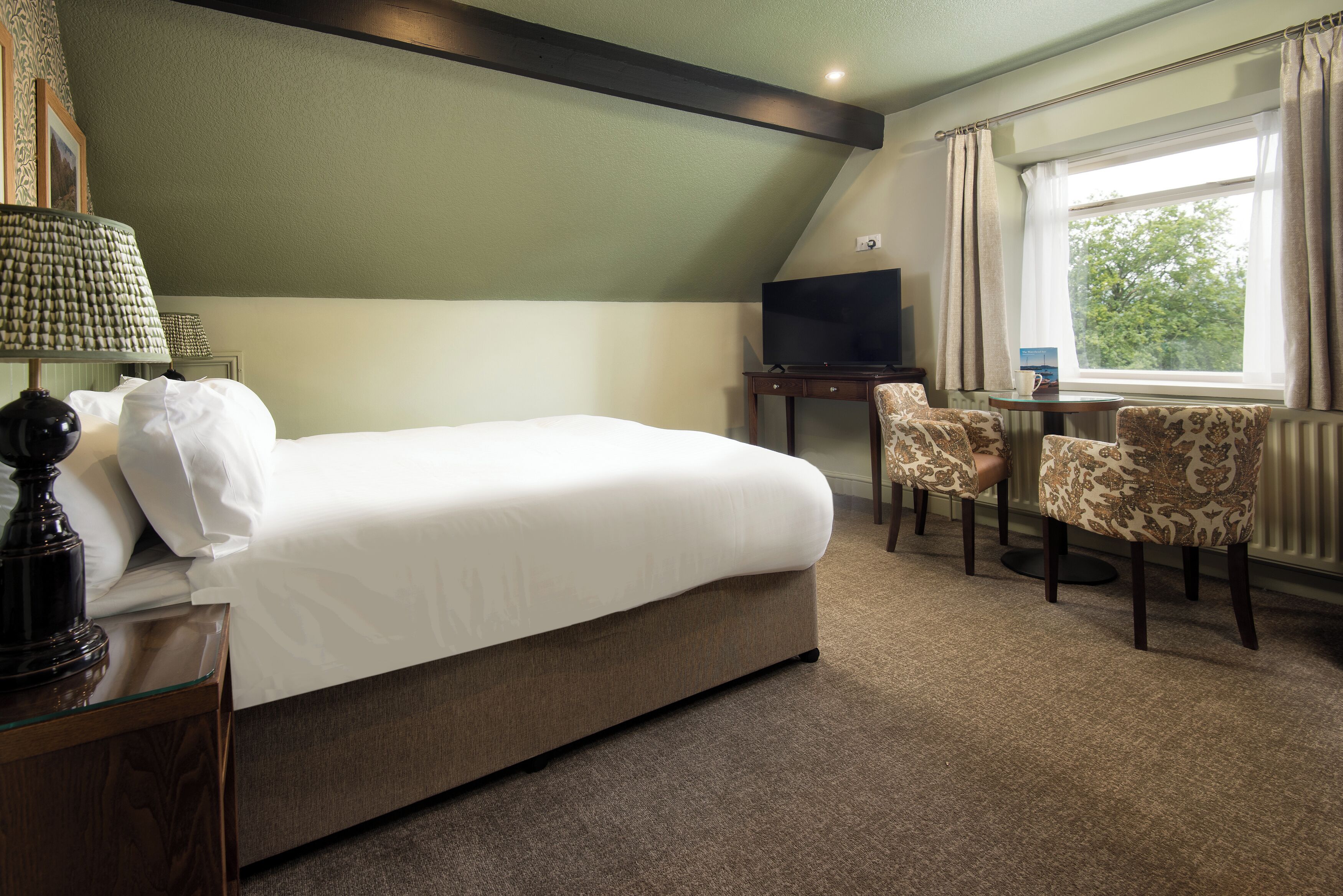 A hotel room with a bed, a desk, two chairs, a TV, and a window with curtains.