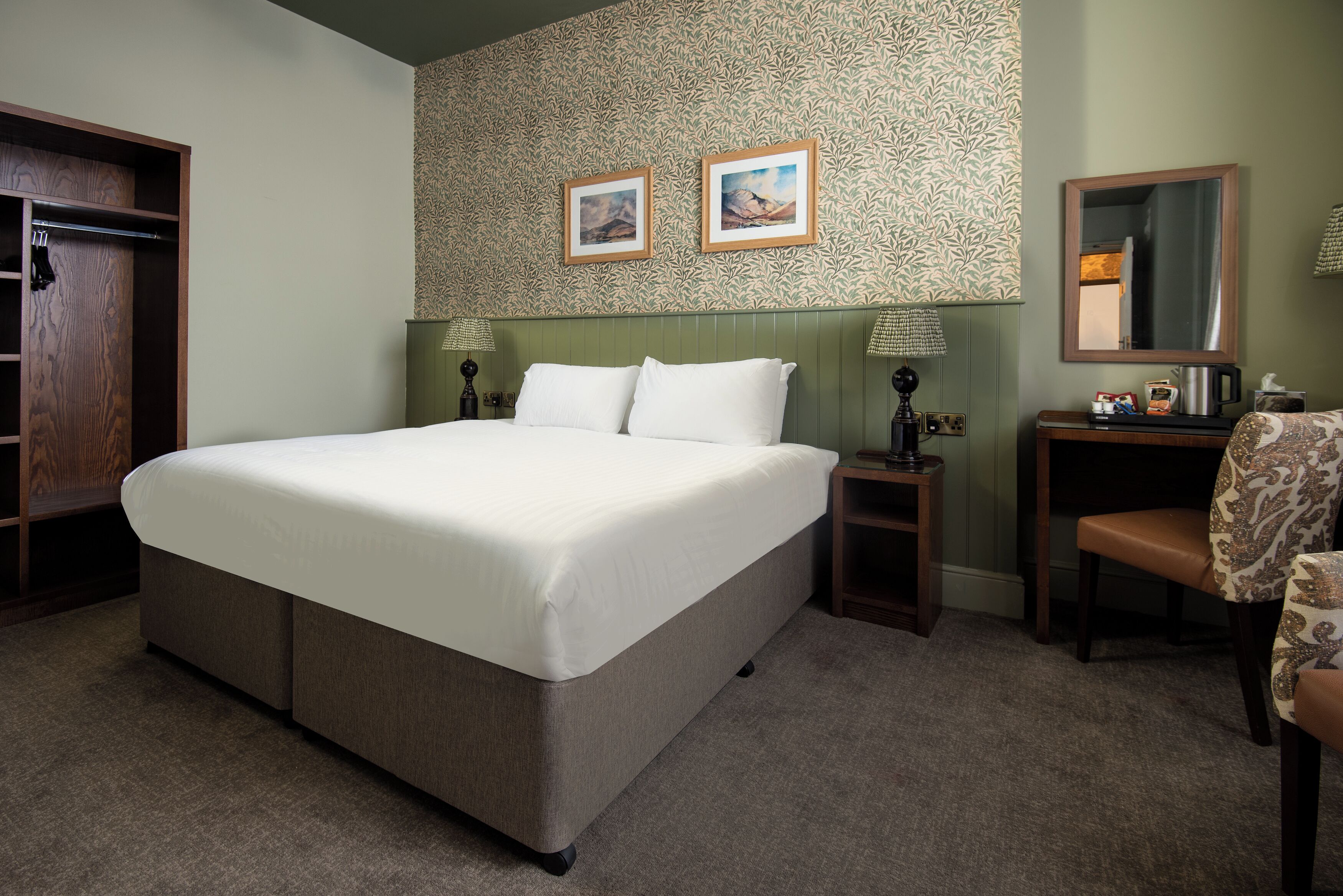 A hotel room with a large bed, a wooden wardrobe, a small table with a lamp, a chair, and two framed pictures on the wall.