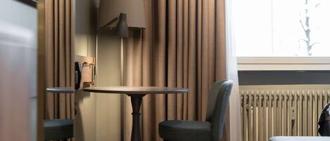 Una habitación con una mesa, dos sillas, una lámpara, un radiador y una ventana con cortinas.