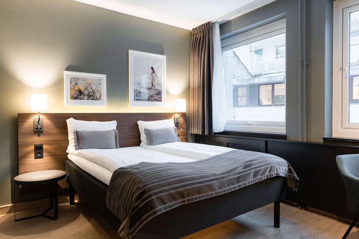 Hotelkamer met een bed, twee wandlampen, een raam met gordijnen en uitzicht op gebouwen.