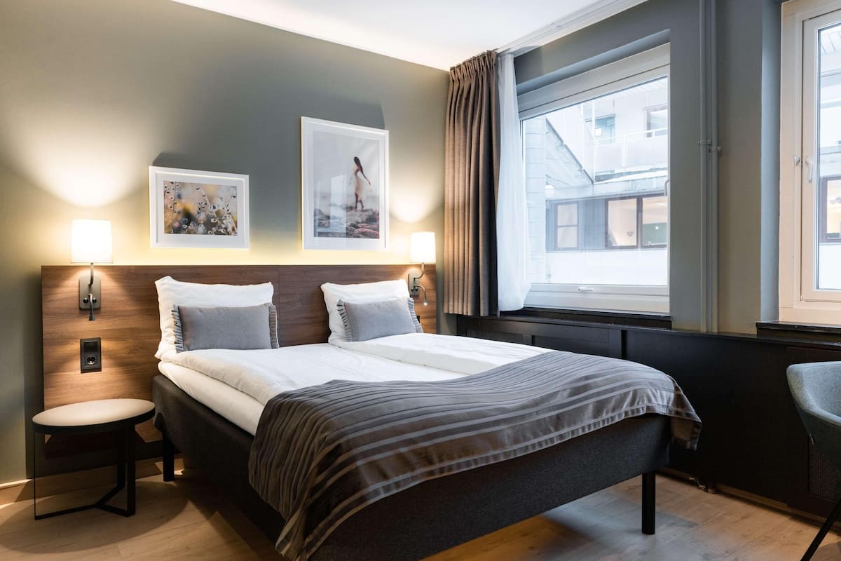A hotel room with a bed, two wall-mounted lamps, a window with curtains, and a view of buildings outside.