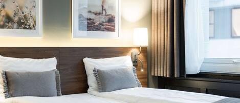 Habitación de hotel con una cama bien hecha, dos cuadros en la pared, una lámpara de noche y una ventana con cortinas.