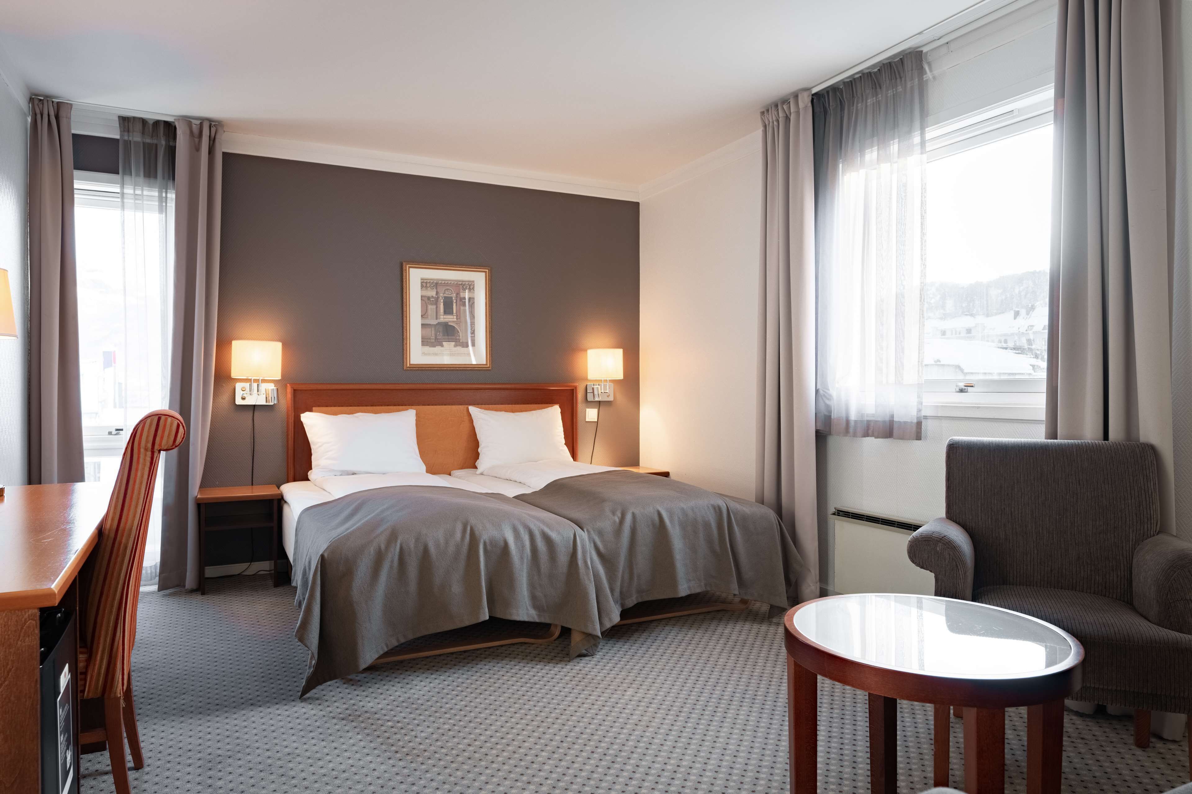A hotel room with a bed, two bedside lamps, a chair, a small table, and a window with curtains.