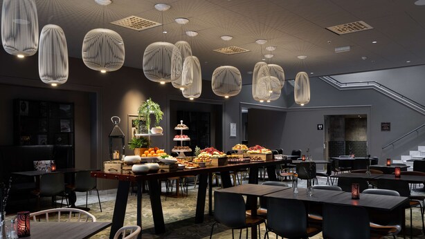A modern dining area with multiple tables and chairs, decorative hanging lights, and a buffet table with various food items. - Scandic Bergen City (Bergen)