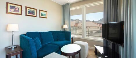 Habitación de hotel con sofá azul, televisor, mesita y ventana con vistas a edificios.