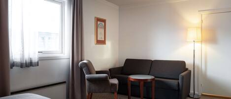 Una habitación de hotel con cama, sillón, sofá, una mesita y una lámpara de pie.