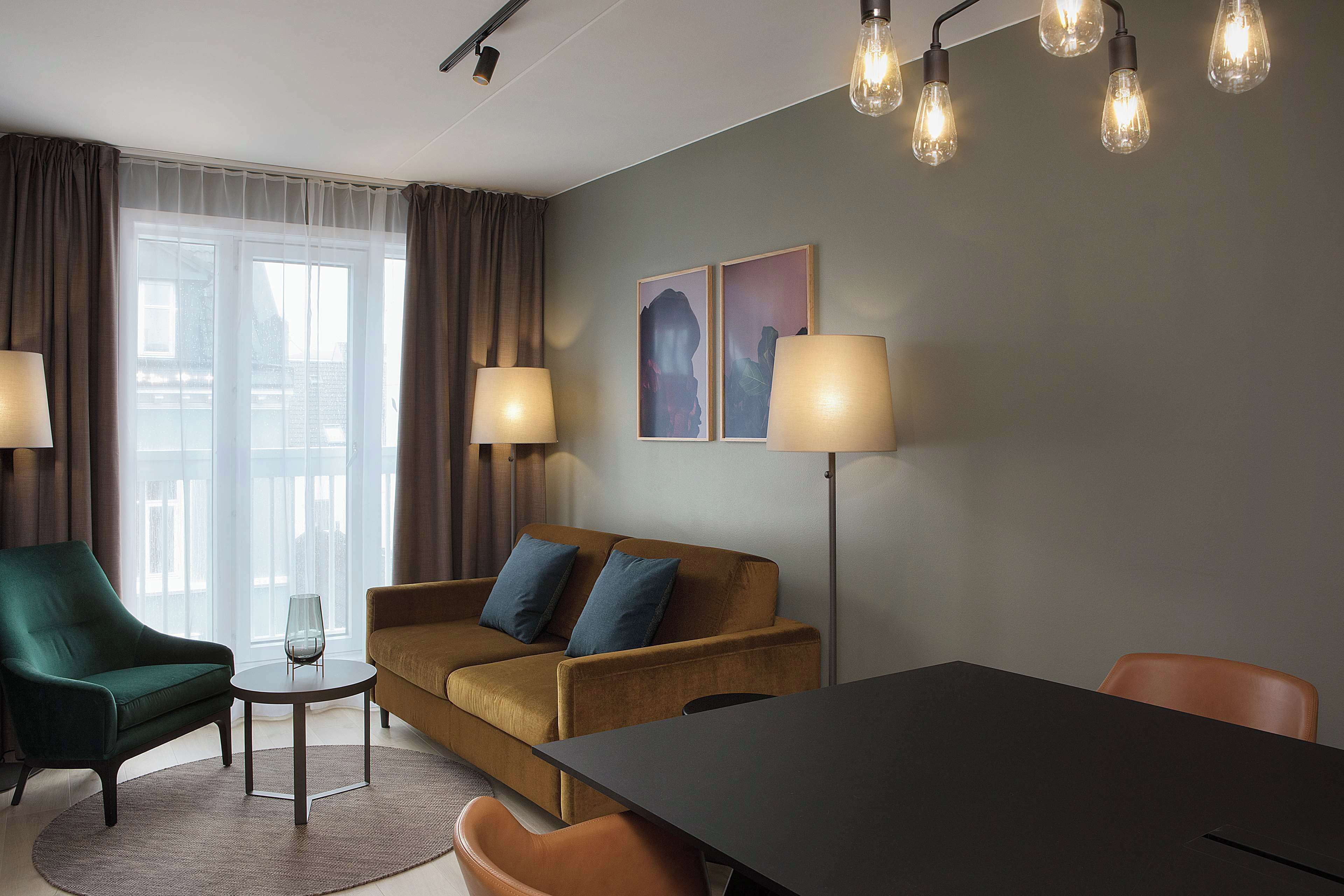 A modern living room with a sofa, armchairs, a coffee table, and a dining table. There are two lamps, a large window with curtains, and artwork on the wall.