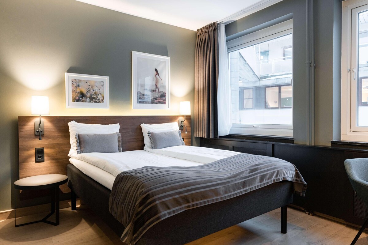 Une chambre d’hôtel avec un lit, deux lampes fixées au mur, une fenêtre avec des rideaux et une vue sur les bâtiments environnants.