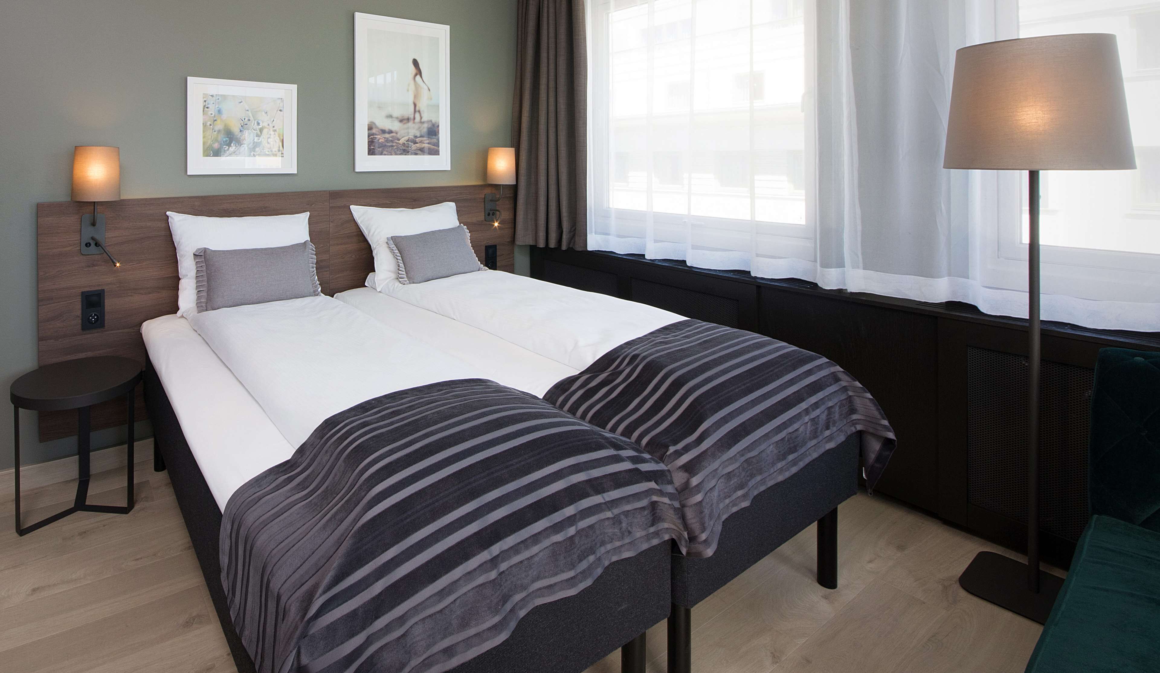 Una cama bien hecha con ropa de cama blanca y mantas a rayas, una mesita de noche con una lámpara, y dos cuadros en la pared.