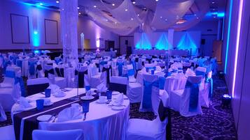 A banquet hall with rows of tables set for a formal event, draped with white and blue linens, and illuminated by blue lighting.