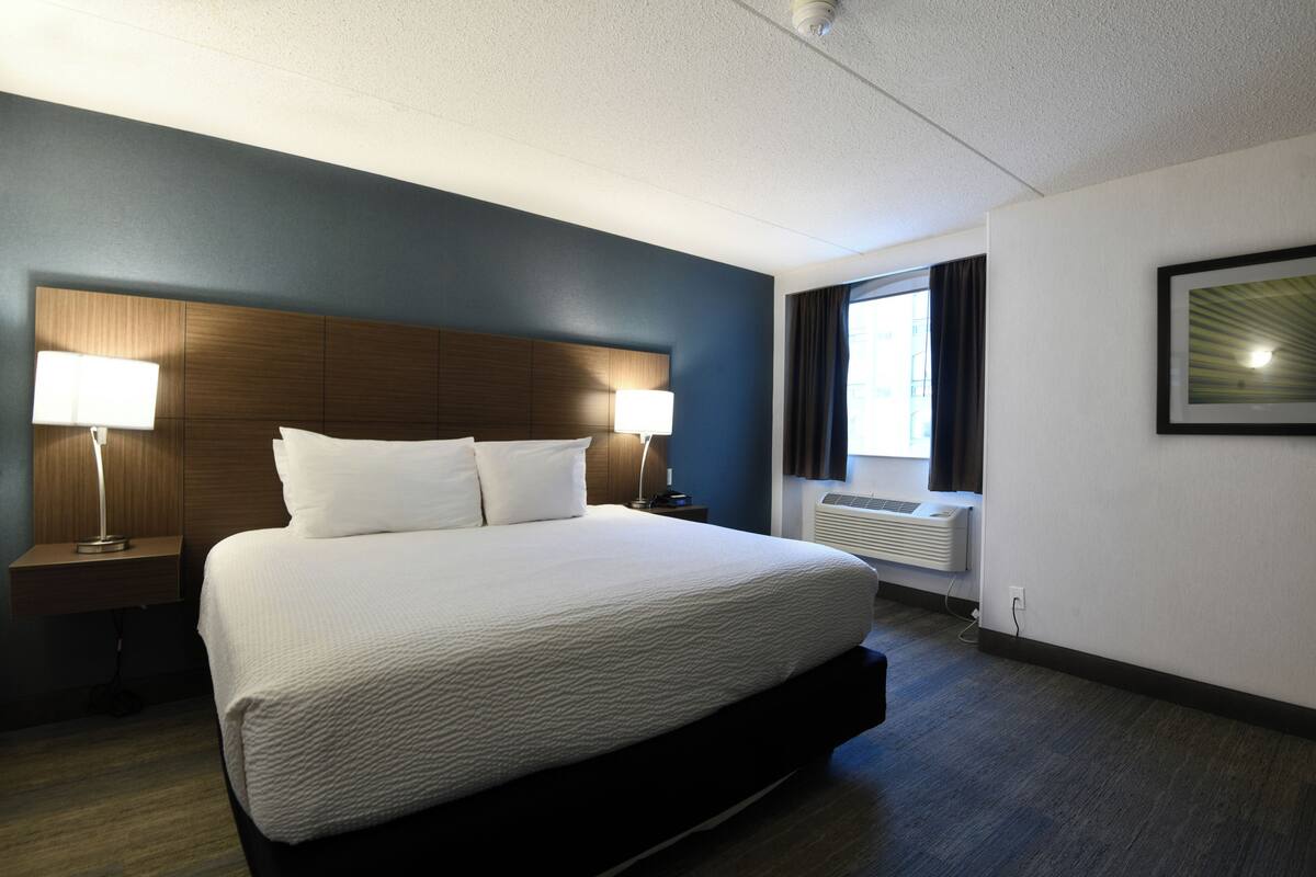 A hotel room with a large bed, two bedside lamps, a window with curtains, and a wall-mounted air conditioner.