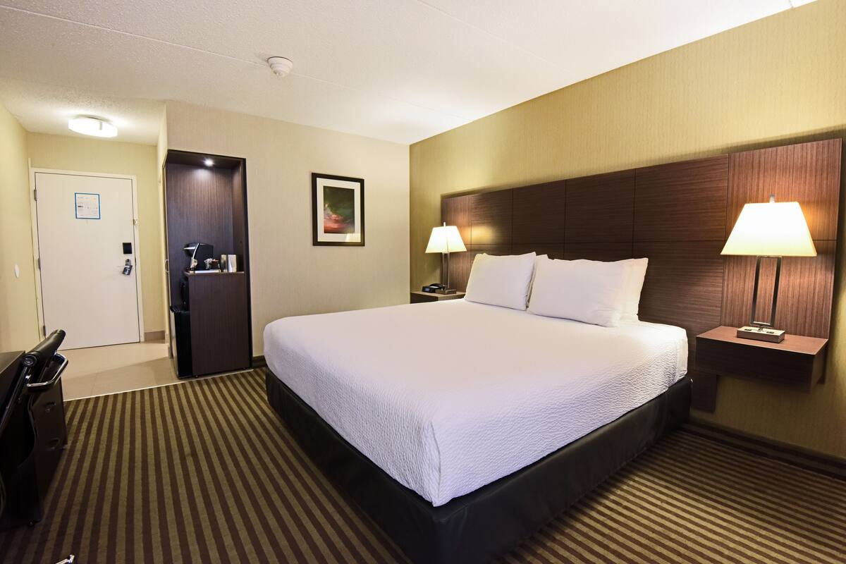 A hotel room with a large bed, two bedside lamps, a desk with a chair, and a door with a sign.