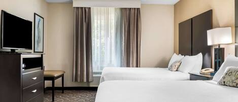 Habitación de hotel con dos camas, un escritorio con televisor, una silla, una lámpara y una ventana con cortinas.