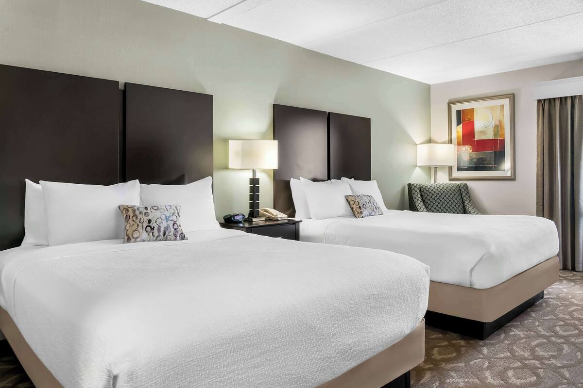 Een hotelkamer met twee bedden, elk voorzien van witte lakens en decoratieve kussens. Er staan nachtkastjes met lampen en een telefoon. Aan de muur hangt een schilderij, en er is een raam met gordijnen te zien.