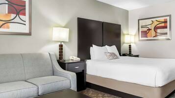 A hotel room with a bed, a grey sofa, a nightstand with a lamp, and artwork on the walls.