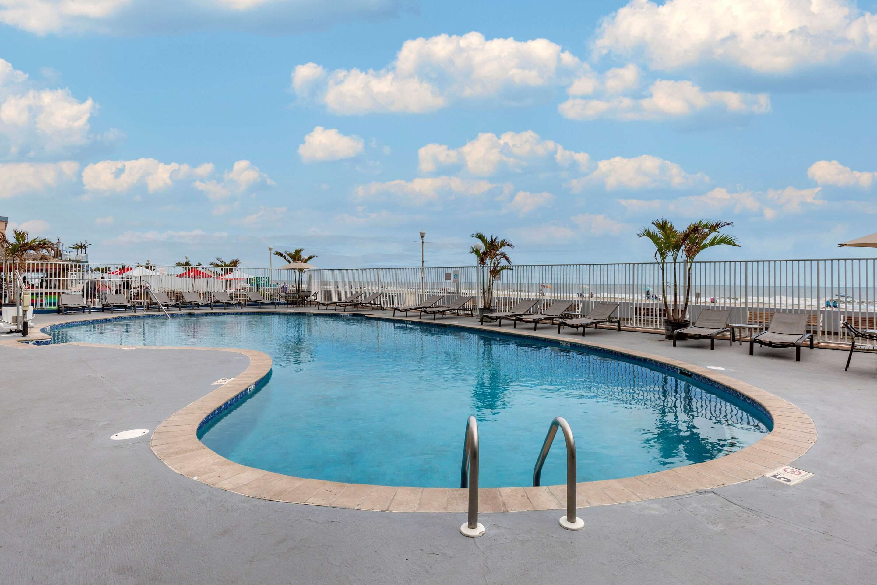 A pool area with a curved design, surrounded by lounge chairs and palm trees, under a clear sky.