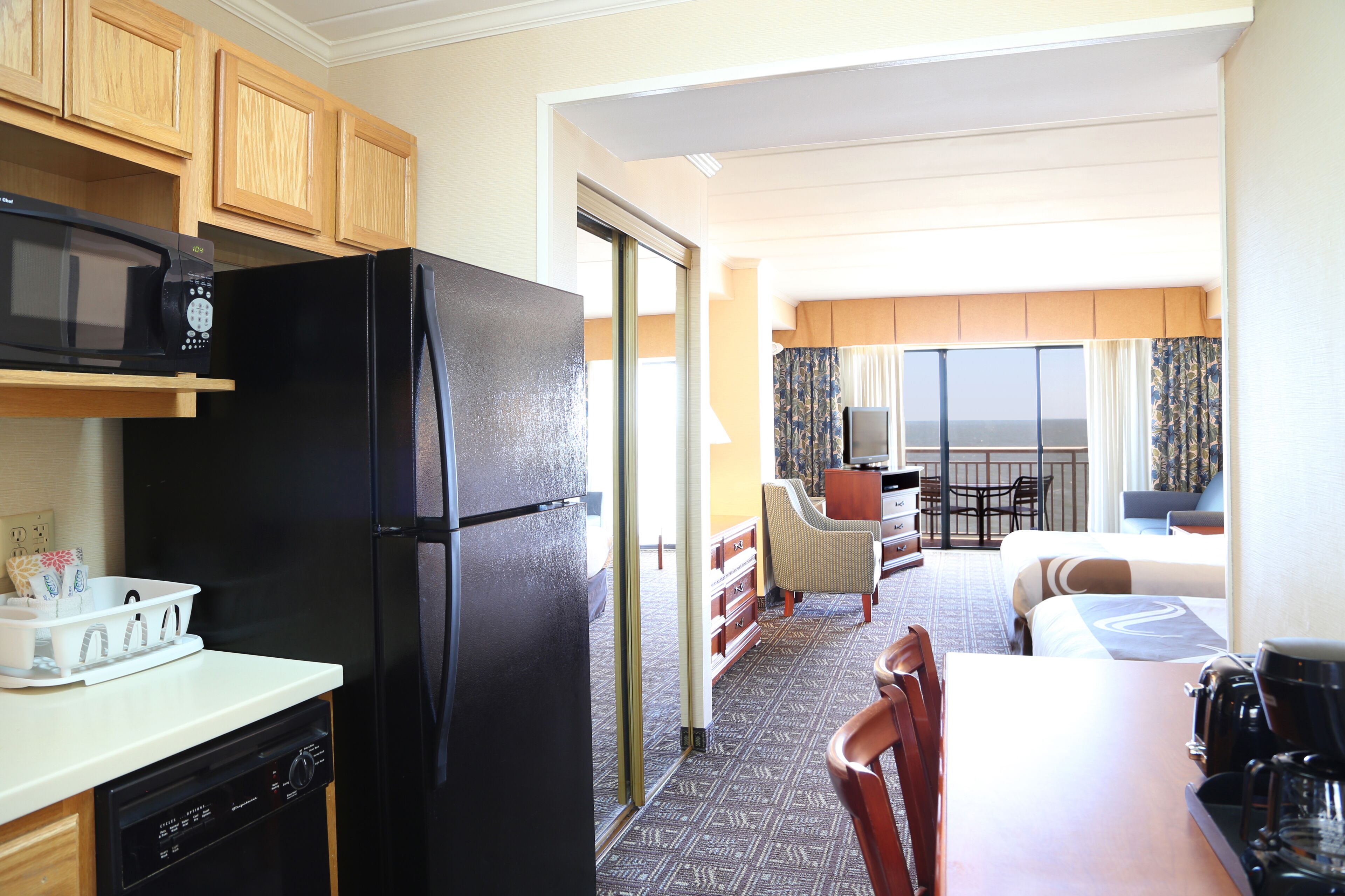 A compact hotel room with a kitchen area featuring a black refrigerator, a microwave, and a dishwasher. The room includes a dining table with chairs, a small sofa, and a balcony with a view.