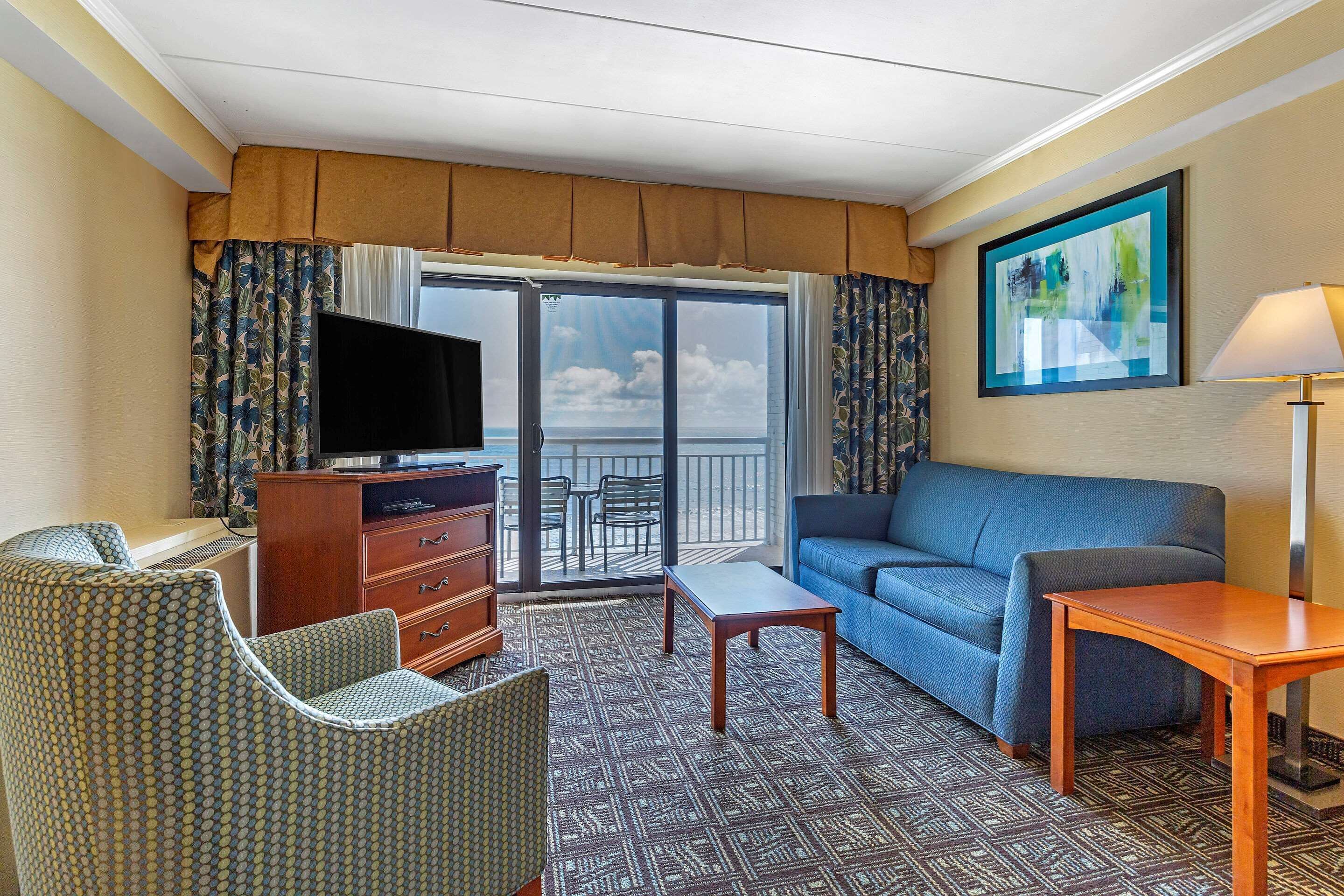A hotel room with a balcony, a sofa, a TV, a desk, and a patterned carpet.